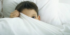 Boy in bed, peeking out behind sheet with one eye